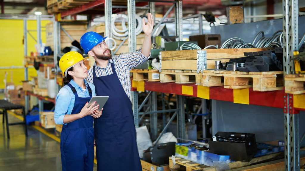 Portrait of two modern factory workers, man and woman, wearing hardhats doing asset and inventory tracking standing by tall shelves in warehouse pointing up